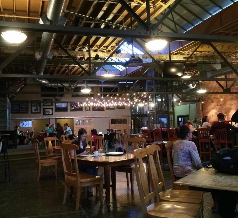 Cozy coffee shop interior with warm lighting, wooden furniture, and patrons working or chatting.