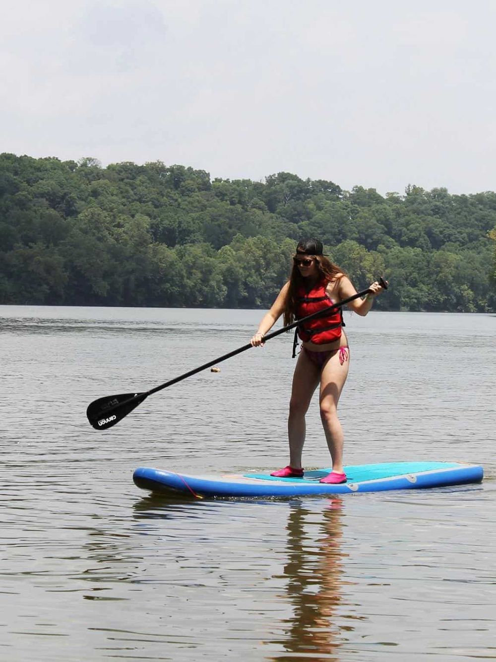 Sedan woman paddleboarding on calm lake with lush green trees in background.