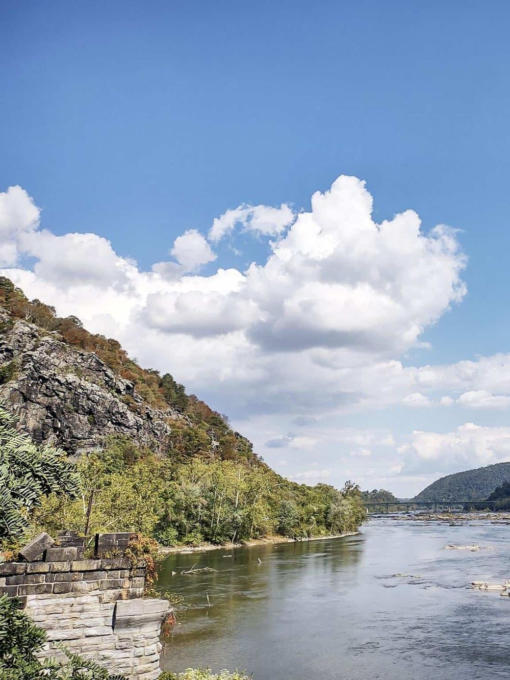 Serene river flowing through lush green landscape with rocky cliffs under blue sky and white clouds.