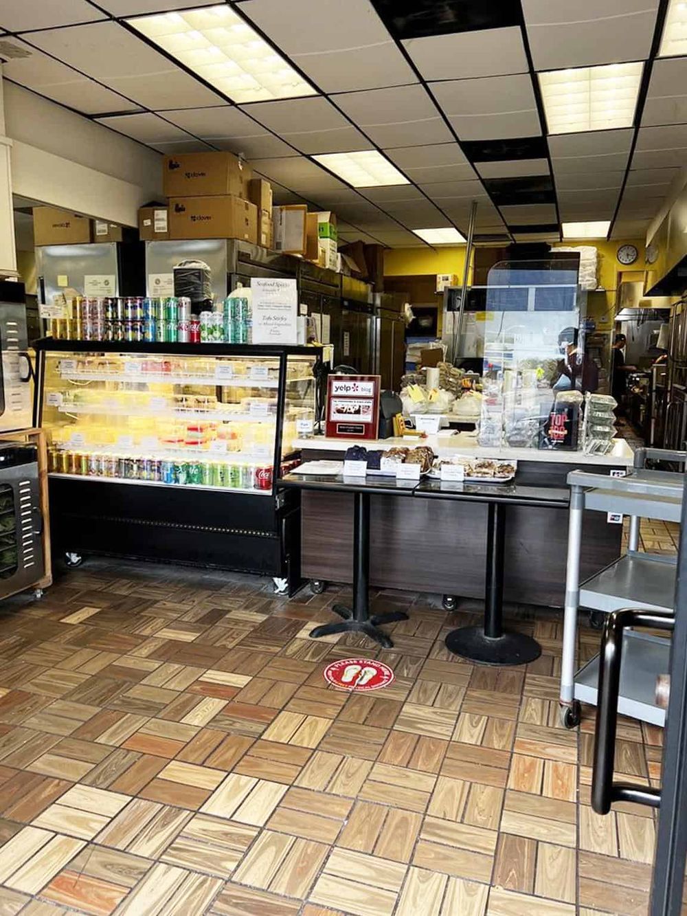 Bright interior shot of a small eatery or deli with snack display, food items, and customers in the background.