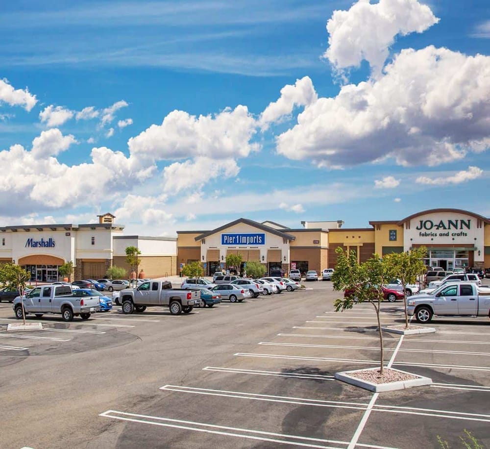 Bright parking lot outside retail stores in shopping center with stores like Pier 1 Imports and JoAnn Fabrics, under a blue sky.