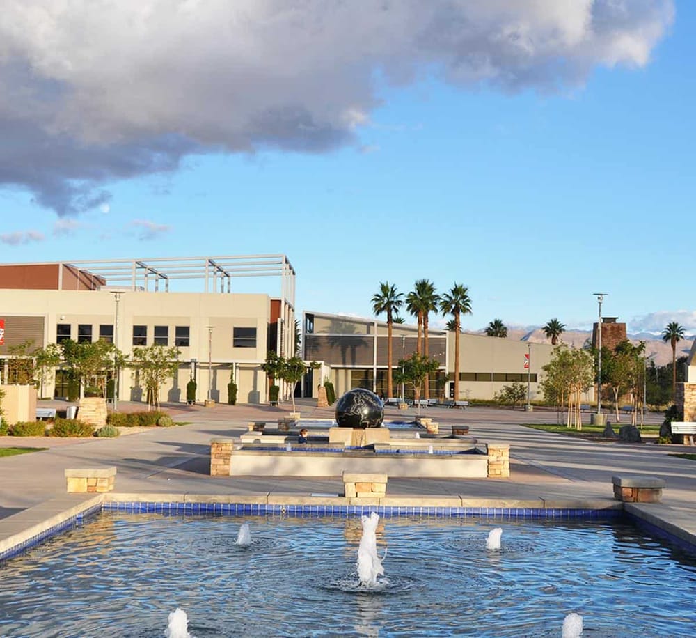 Modern shopping mall with palm trees, water fountain, and mountain backdrop in sunny weather.