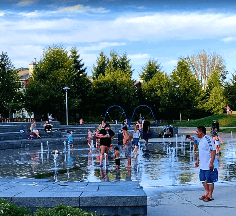 Spray park water feature with children playing and families enjoying a sunny day, outdoor fun in a park setting.