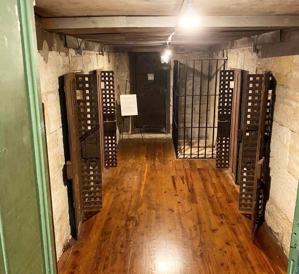Old wine cellar with wooden racks and metal bunk, stone walls, and wooden floor.