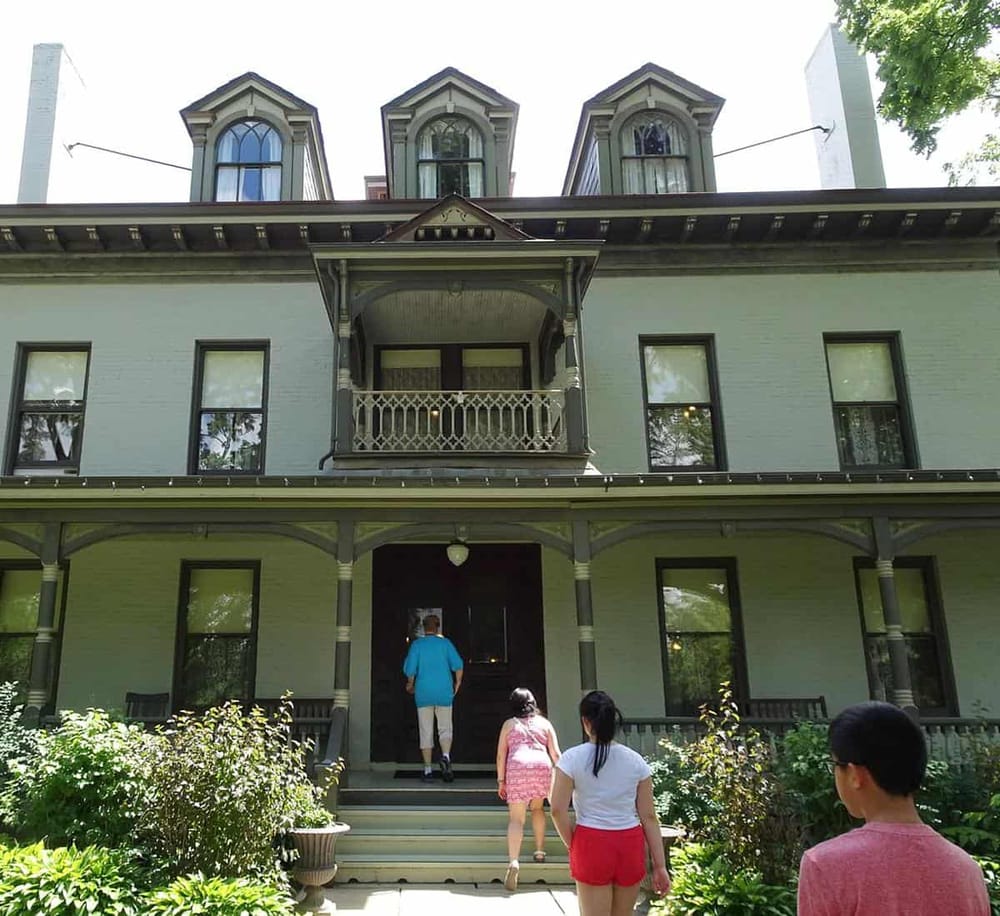 Victorian-style historic house with front porch, located in a lush green setting.