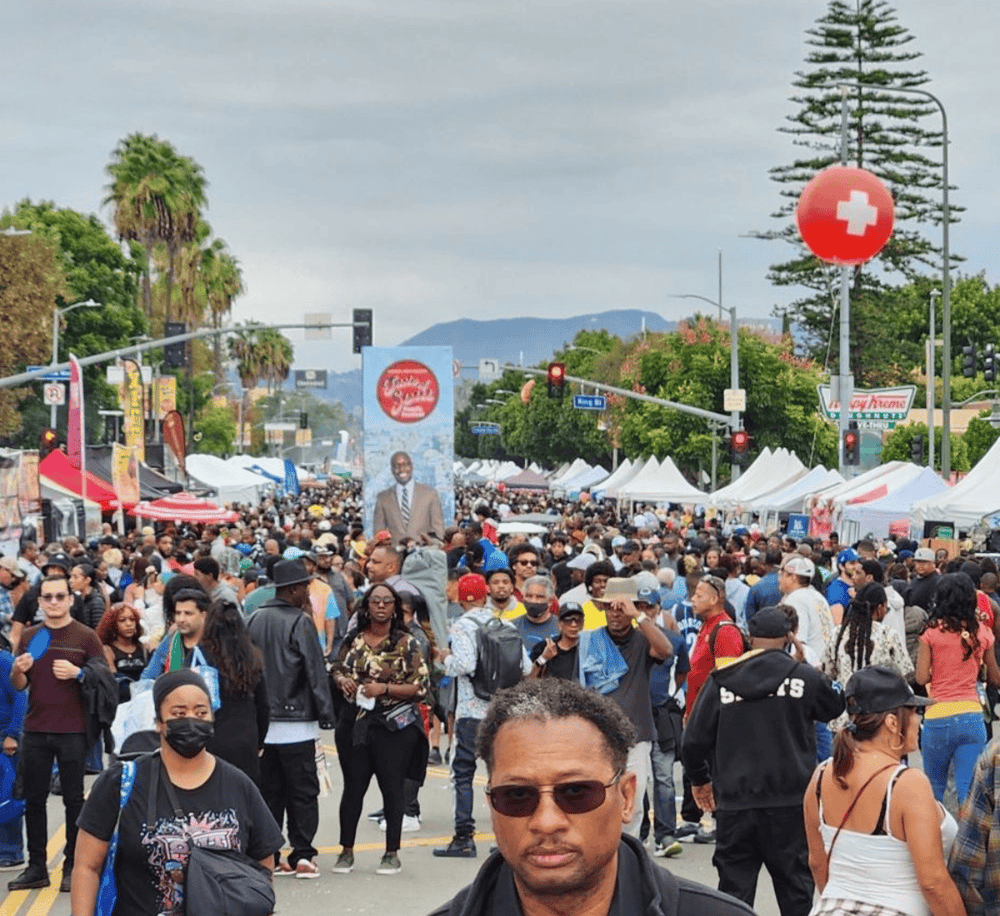 Crowd at outdoor event with tents, banners, and street signs, showcasing community gathering in an urban setting.