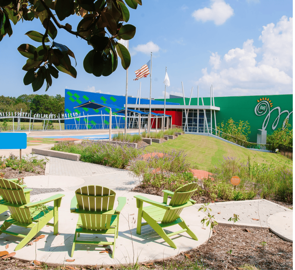 Bright colorful children's museum exterior with outdoor seating, lush landscaping, and American flags.