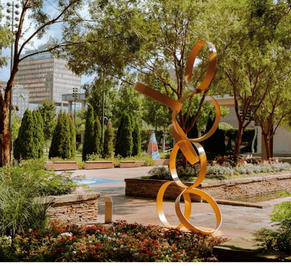 Modern outdoor sculpture in downtown park with lush greenery and city buildings in background.