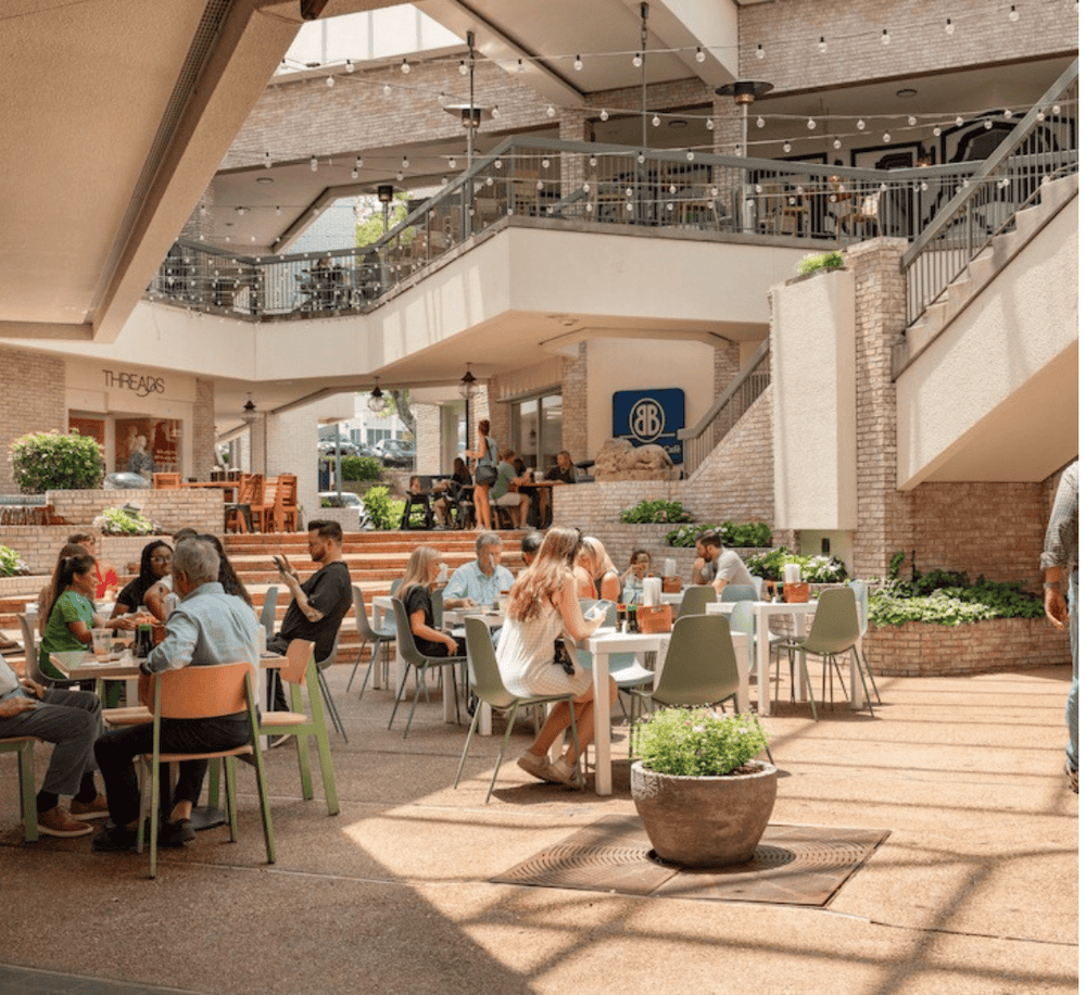 Alto text only (max 100 characters): Outdoor dining at QuestForDirections shopping mall with people enjoying sunny day.