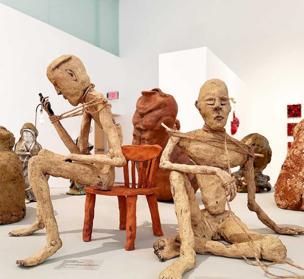 Fragile humanoid sculptures made of clay, depicting seated figures with rope bindings, displayed in an art gallery.