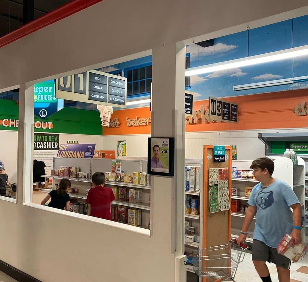 Bright grocery store aisle with children shopping and shelves filled with cereal boxes and snacks, under store signage.