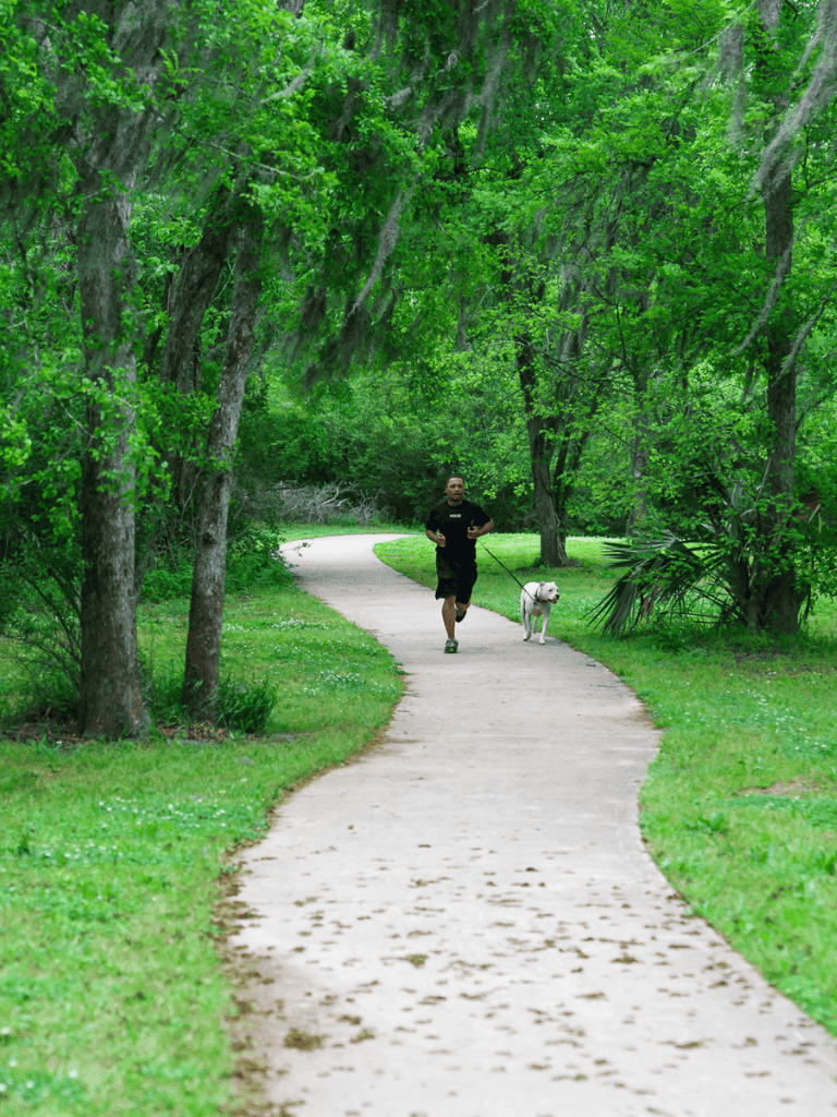 Man jogging with dog on paved trail through lush green park, outdoor activity, fitness, nature walk.