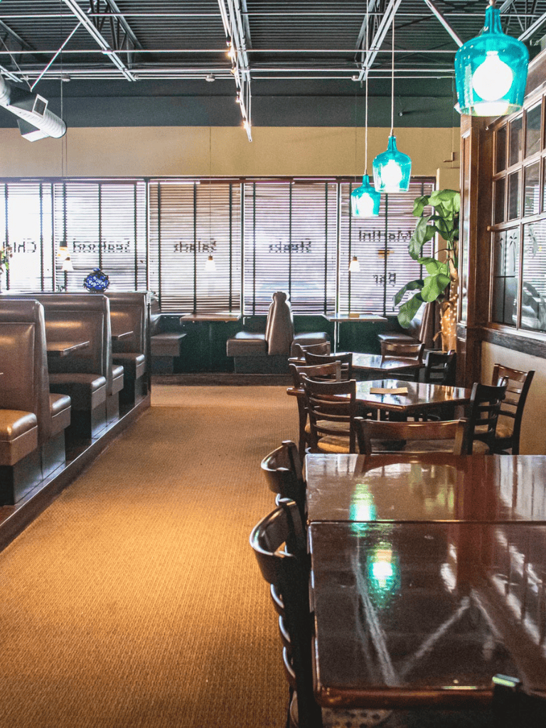 Cozy restaurant interior with wooden tables, chairs, booth seating, modern pendant lighting, and large windows.