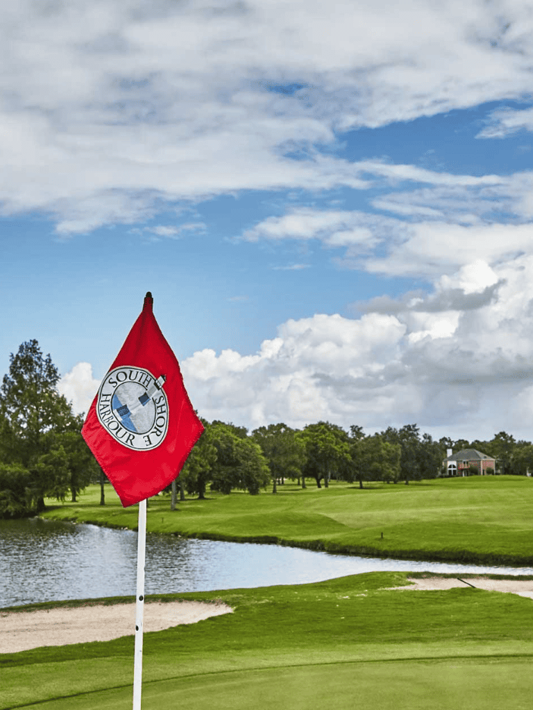 High school golf flag near water hazard on a lush golf course with trees and club house in background.