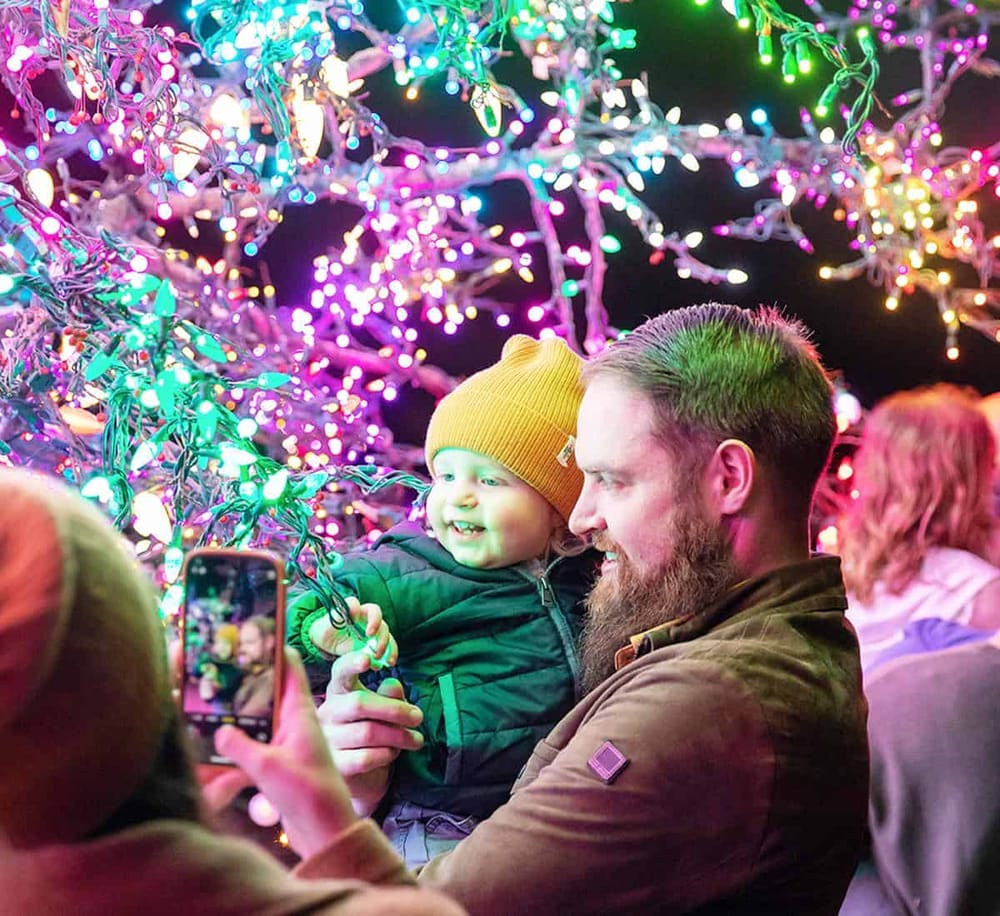 Colorful holiday lights illuminate a joyful moment between a father and son at a festive event.