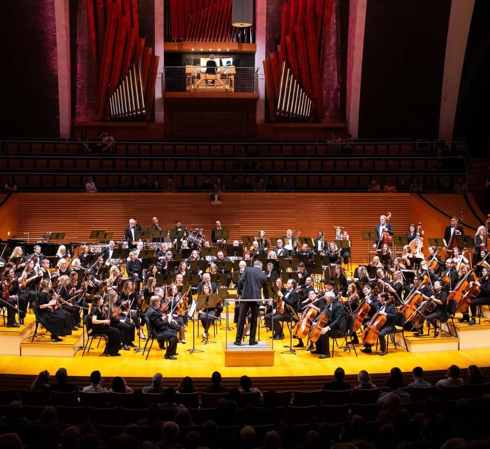 Orchestra performing in a concert hall with an attentive audience and a musical conductor at the center.