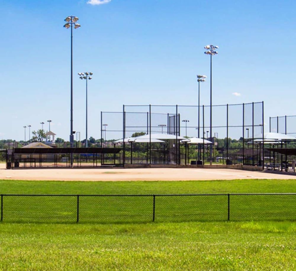 Bright softball field with outfield fencing, stadium lighting, and clear blue sky in the background.