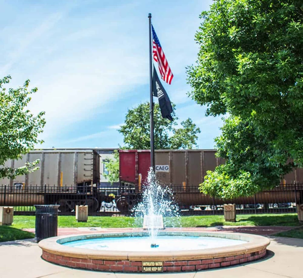 American flags flying near train cars at Quest For Directions train station, outdoor fountain and greenery.