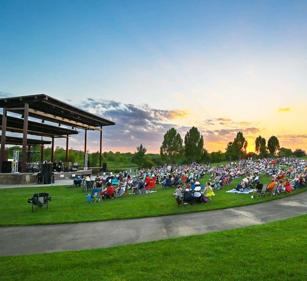 Outdoor concert at sunset with large audience on grass and stage, highlighting entertainment and community event.