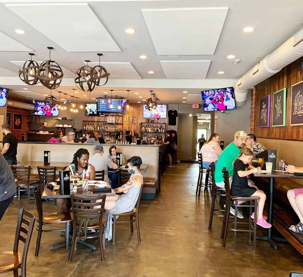 Modern American restaurant interior with guests dining and multiple TV screens, warm lighting, and wood accents.