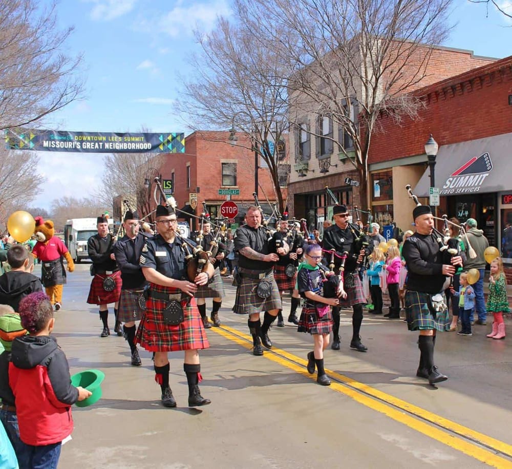 Traditional Scottish pipe band marching during a parade in downtown Lee's Summit, Missouri, with spectators enjoying the event.