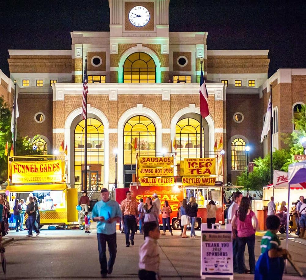 Brightly lit fair at night outside a historic courthouse, featuring food stalls and crowds.