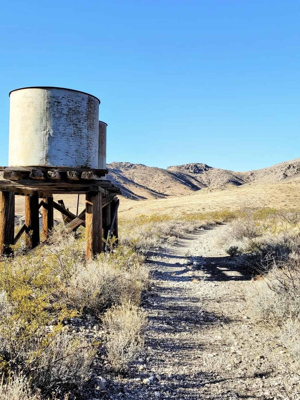 Rustic water tower in desert landscape, hiking trail, dry shrubs, mountains, sunny day, QuestForDirections.