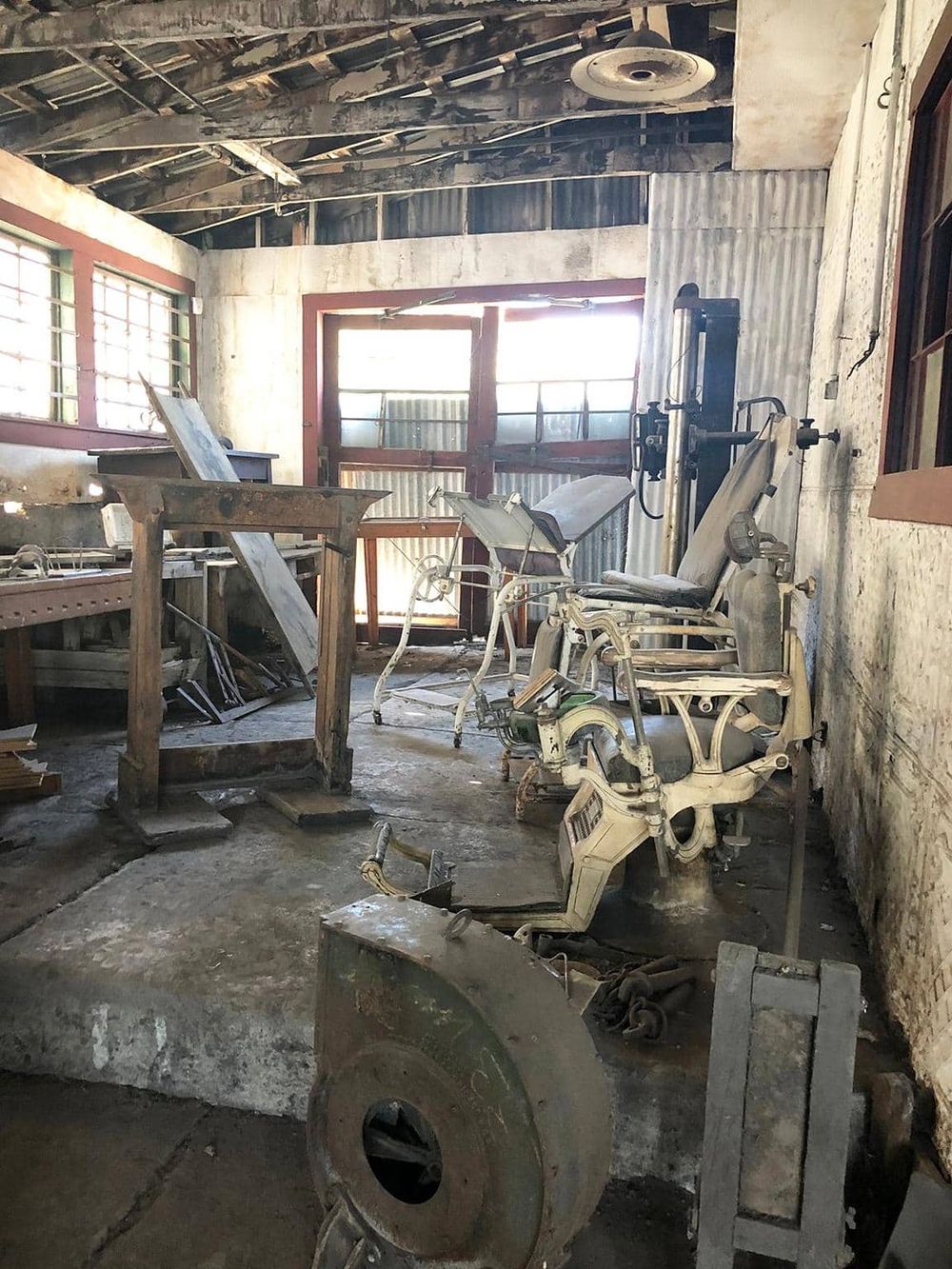 Old medical equipment in a dilapidated hospital room with broken windows and exposed ceiling beams.