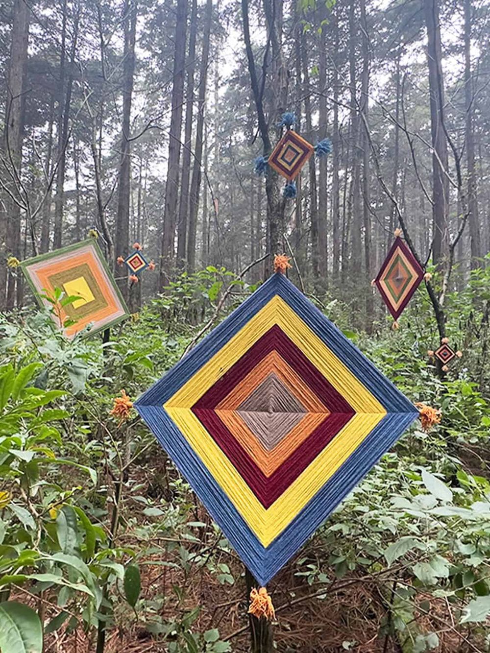 Vibrant colorful diamond-shaped yarn art hanging in a forest setting, creating a whimsical outdoor decor.