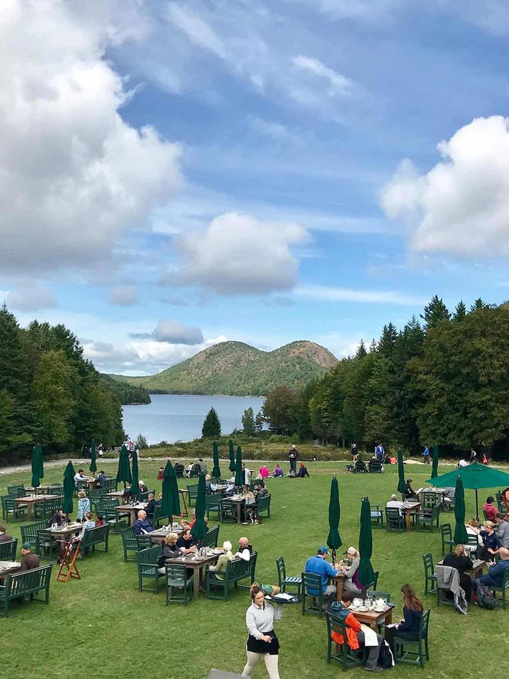 Vibrant outdoor dining area near a lake with lush trees, mountains, and a blue sky, perfect for scenic meals and nature relaxation.