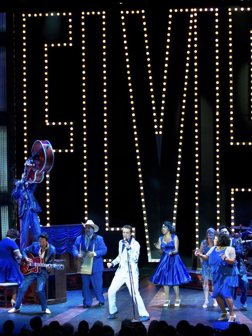 Bright stage with musical performers and large illuminated "ELVIS" sign, lively entertainment scene.