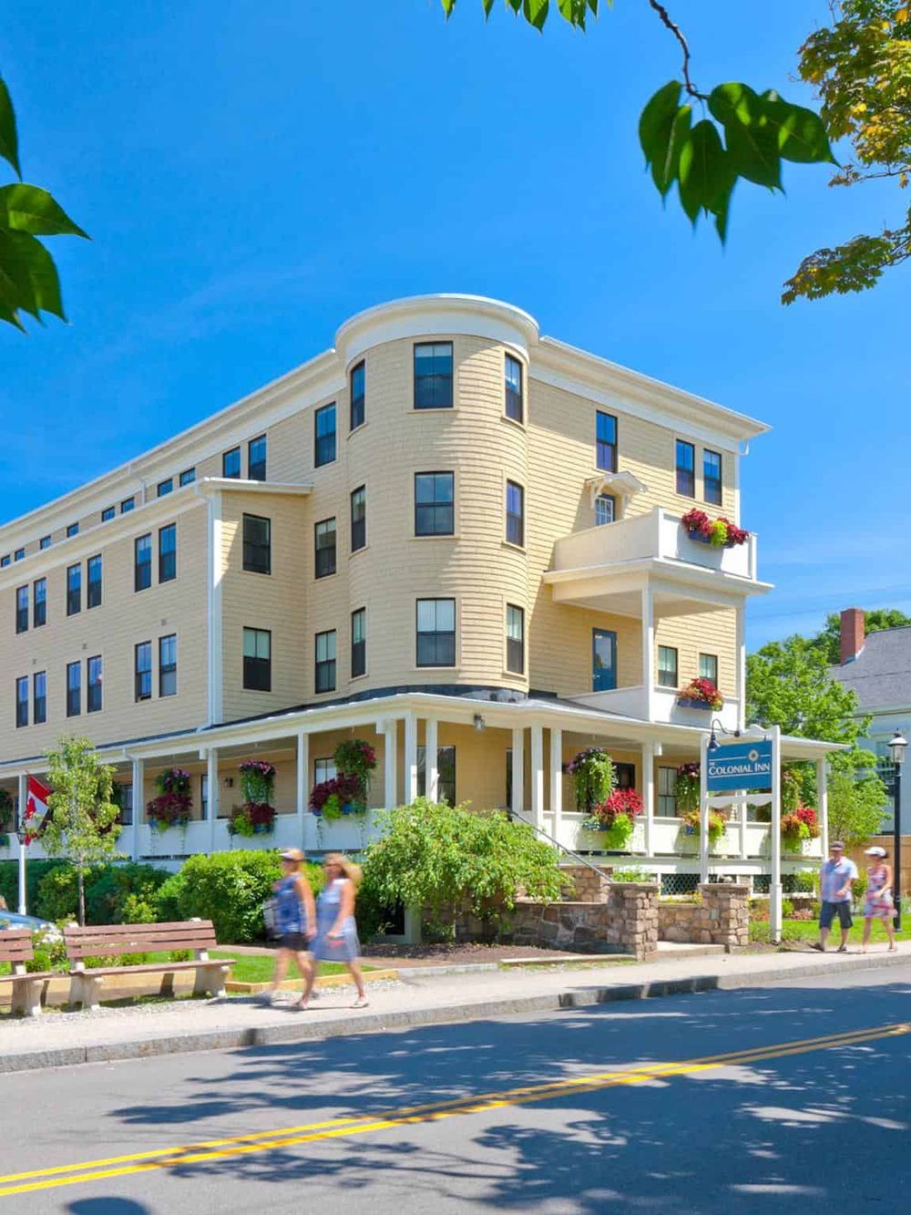 Charming yellow hotel building with colorful flower boxes and people walking, situated on a lively street.