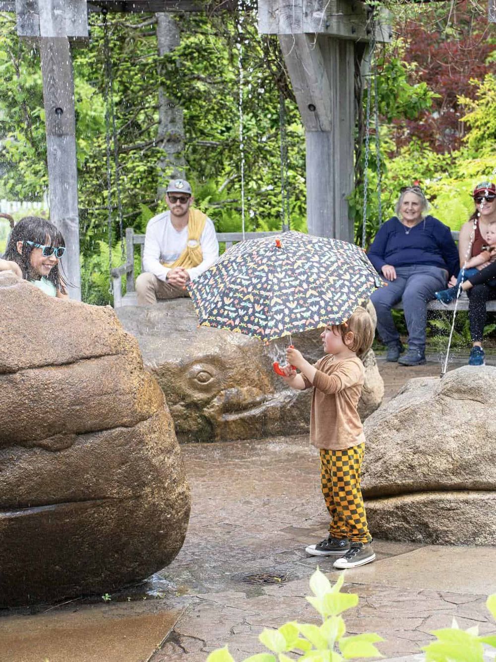 Colorful outdoor scene with children and adults at a fountain, enjoying nature and family moments.