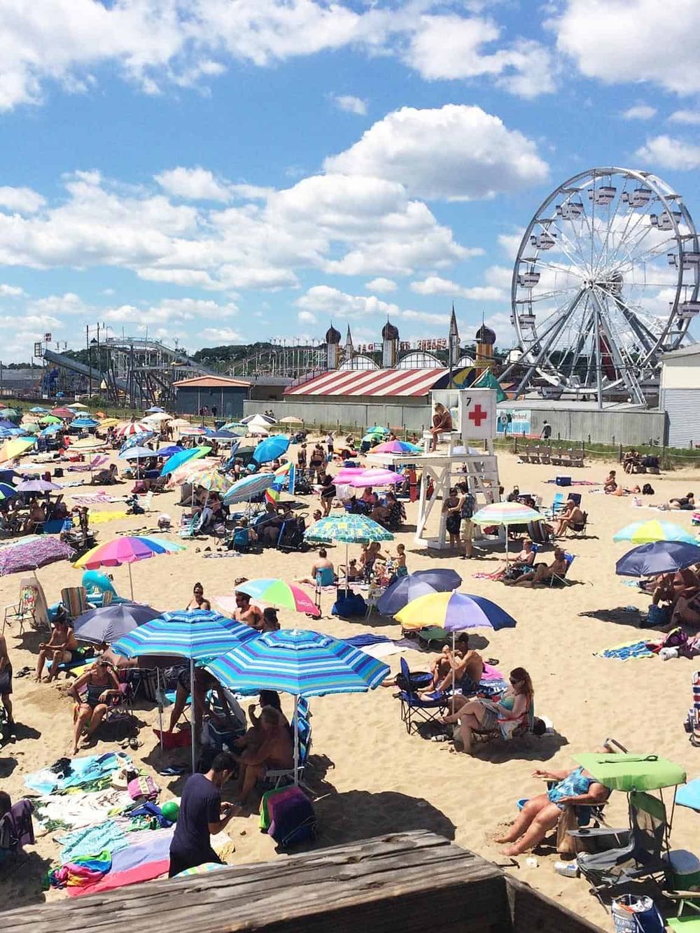 Colorful crowded beach scene with umbrellas and amusement park rides in the background, sunny summer day.