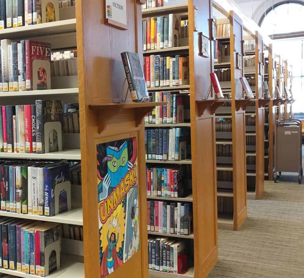 Colorful superhero comic book poster displayed in a public library aisle filled with books.