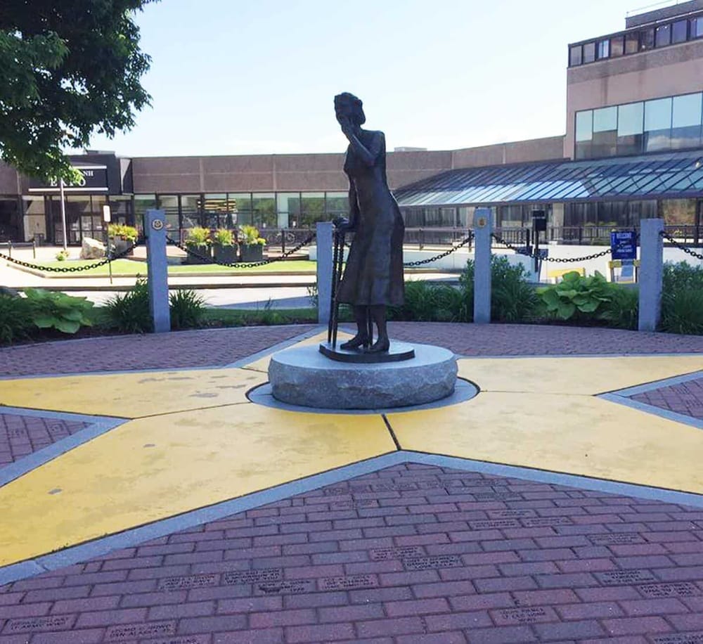 Peaceful outdoor sculpture of a woman with a cane on a circular stone base in a modern urban plaza, surrounded by greenery and chains.