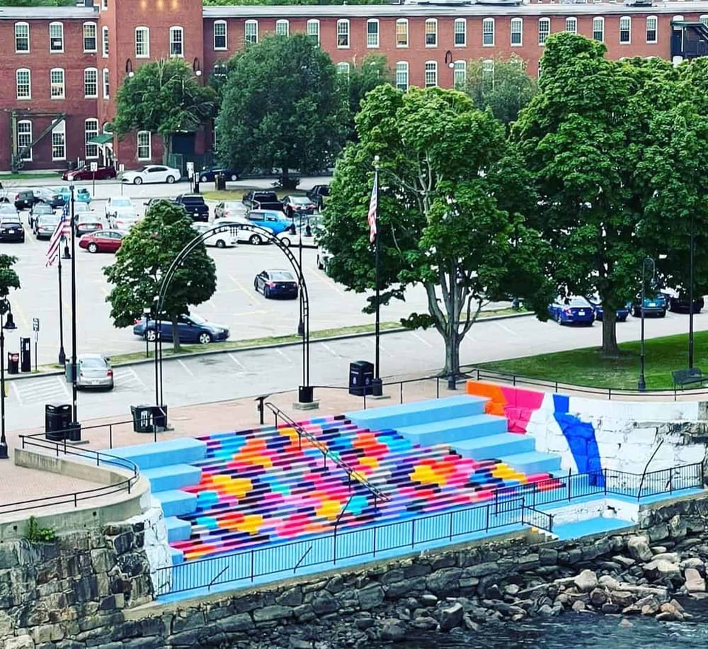 Vibrant colorful staircase near water with trees and parking lot in background, urban outdoor scene.