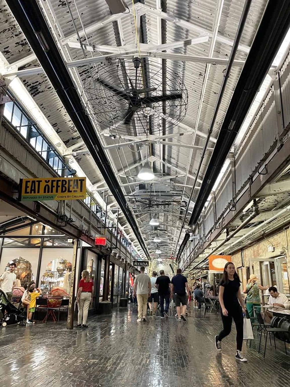 Indoor market corridor with shoppers and ceiling fans, vibrant marketplace atmosphere.