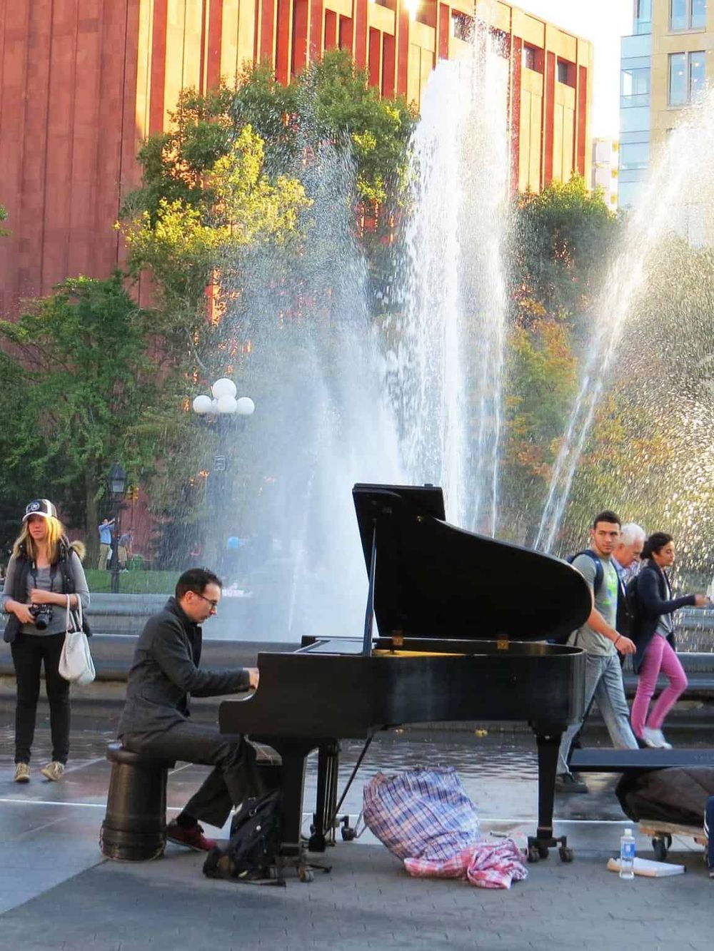 Pianist playing piano outdoors by city fountain, urban park scene, live street performance, people enjoying city life.