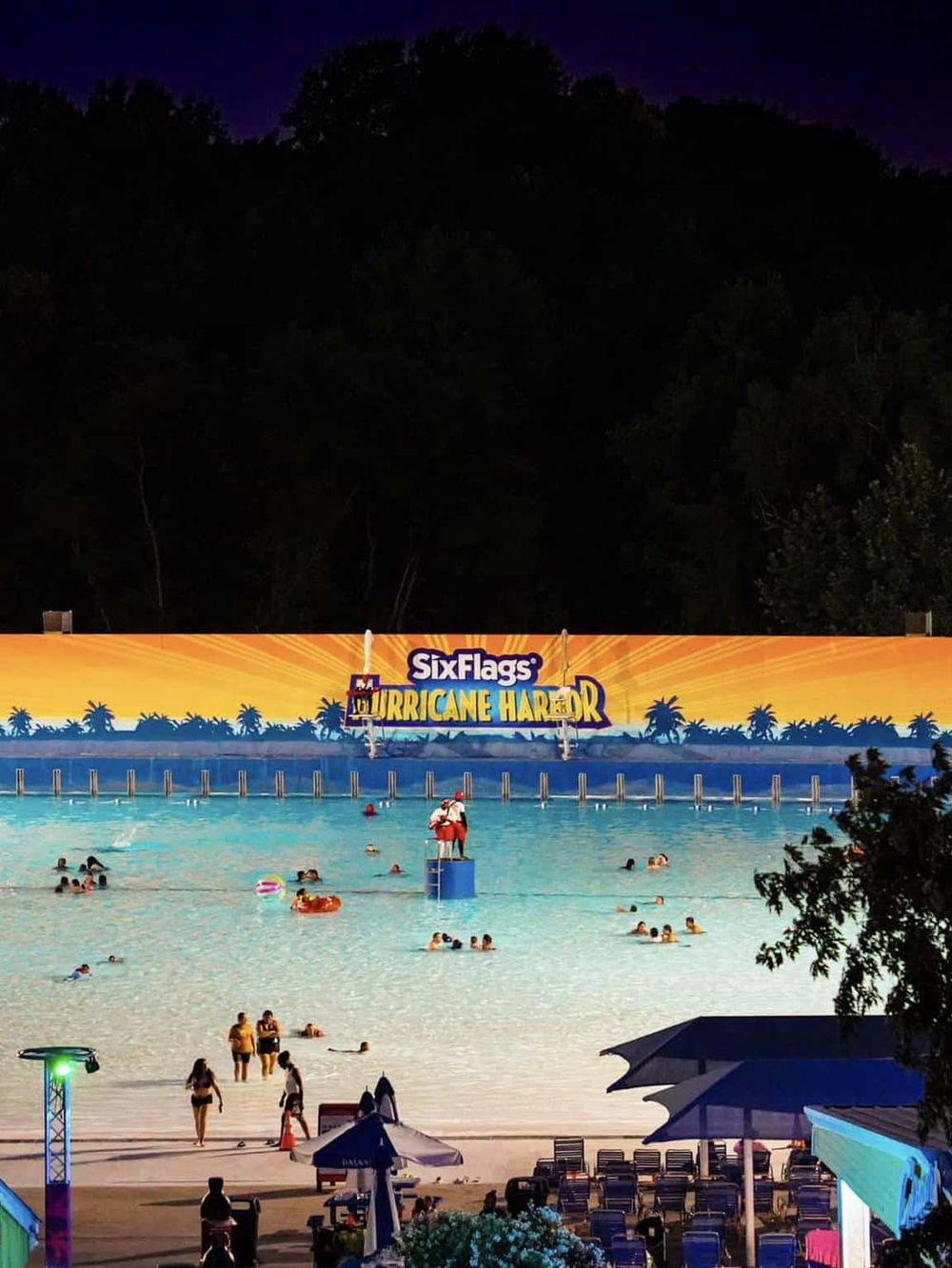 Nighttime scene at a water park with lit-up sign “Six Flags Hurricane Harbor” and people enjoying the water.