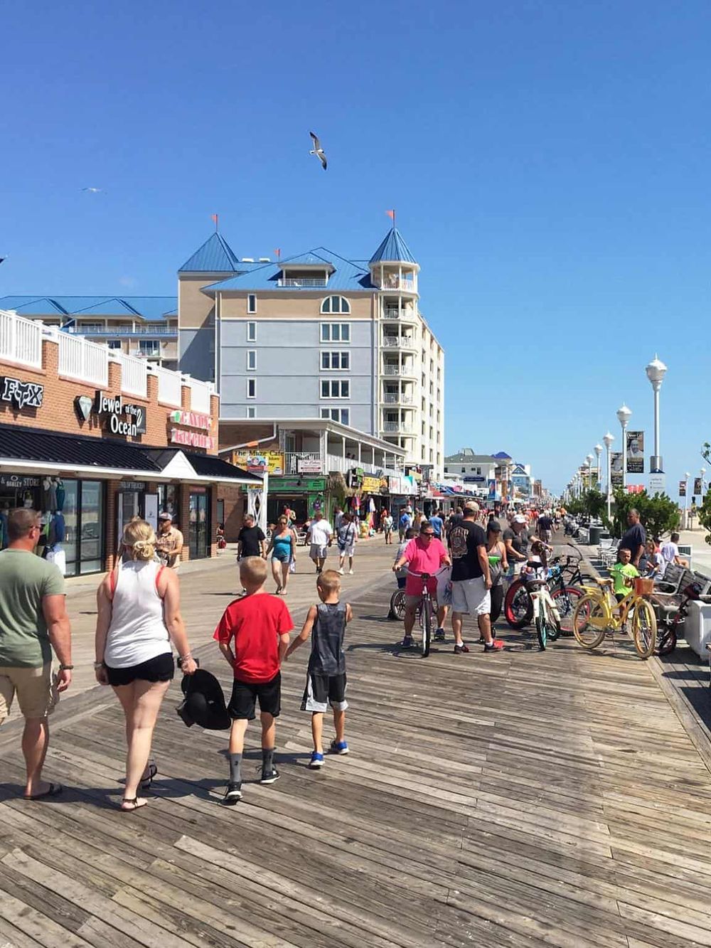 Seaside boardwalk with people, shops, and bicycles under a clear blue sky in a coastal town.