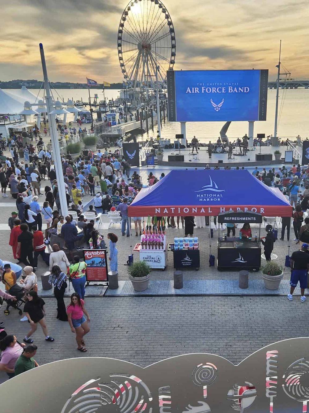 Elevated view of National Harbor waterfront with an outdoor concert, ferris wheel, and crowd enjoying a live event.
