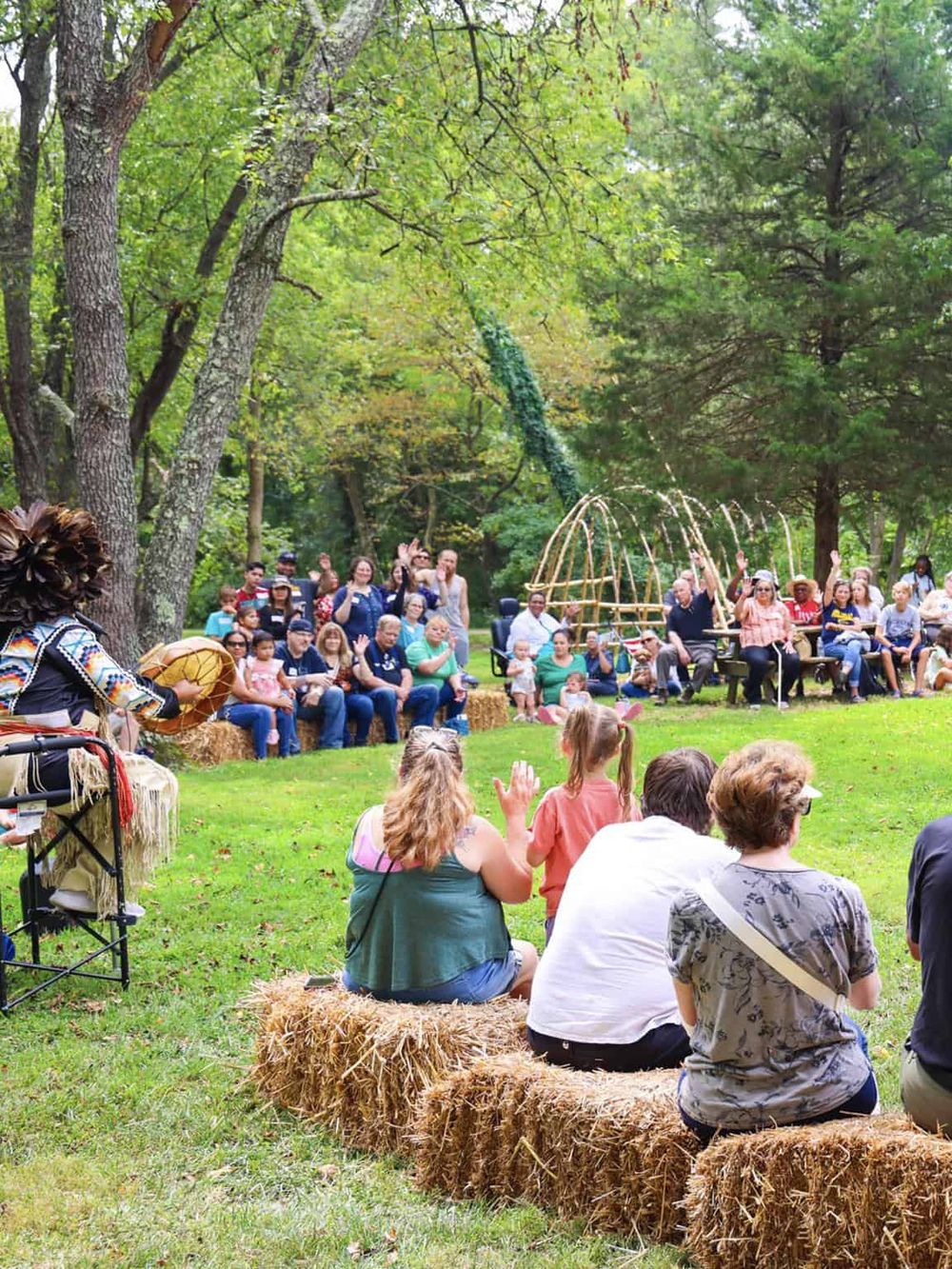 Children and families enjoying outdoor storytime at a rural park with hay bales, surrounded by tall trees.
