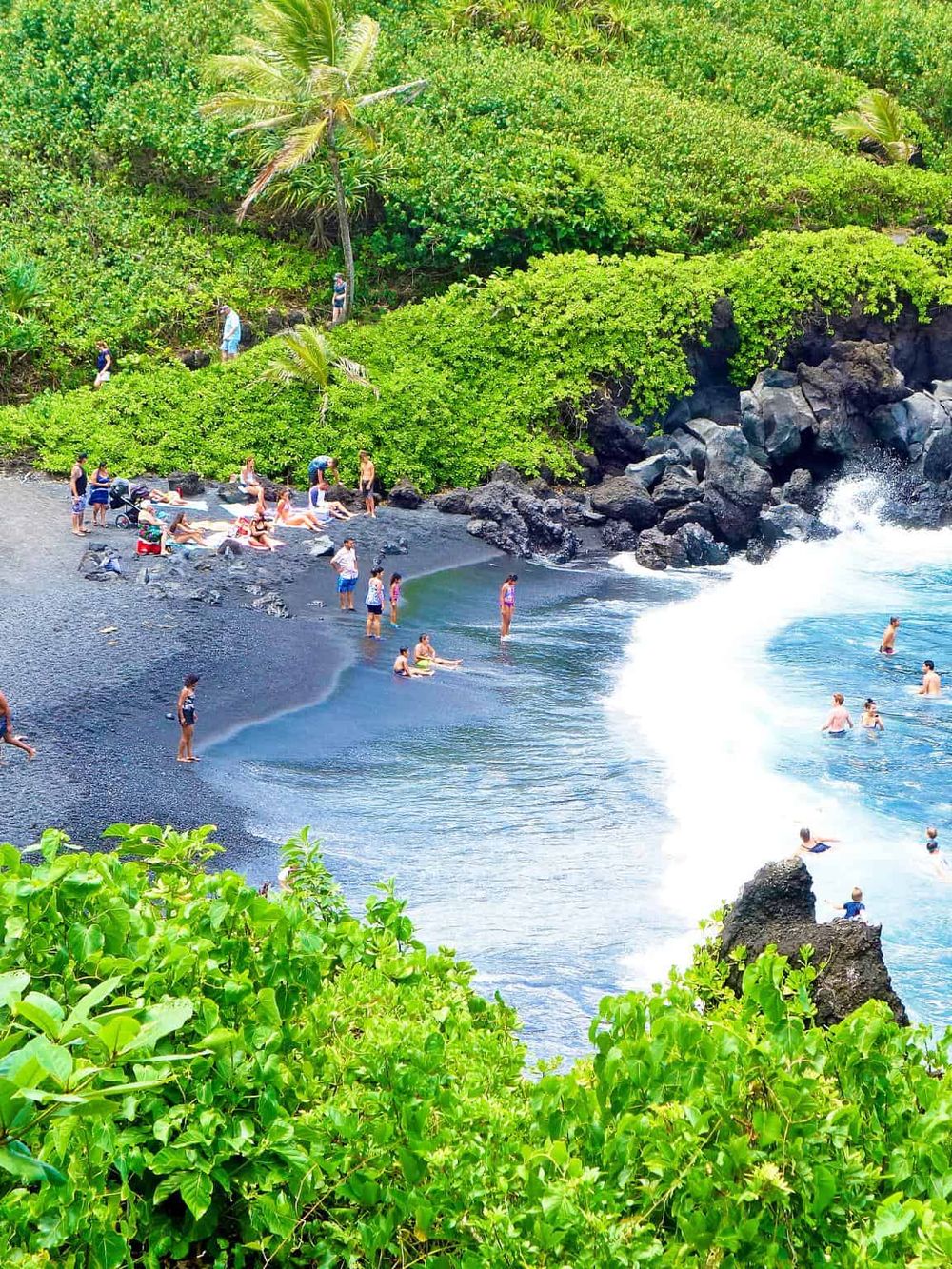 Vibrant black sand beach with sunbathers and surfers, lush green tropical vegetation, and volcanic rocks in Hawaii.