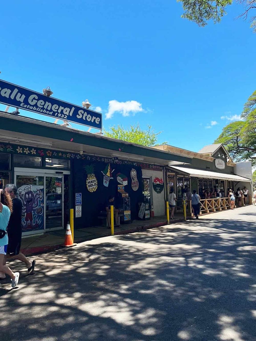 Colorful local store on a sunny day with people in line, outdoor shadows, and clear blue sky.