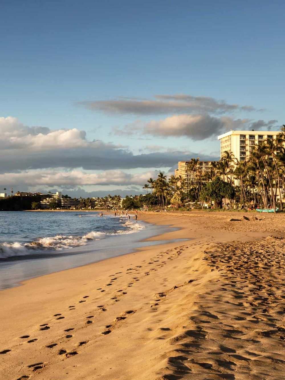 Scenic beach with soft sand, footprints, and ocean waves near high-rise buildings and palm trees at sunset.