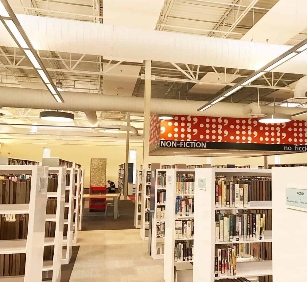 Bright library aisle with bookshelves, sign for non-fiction section, modern lighting, and ceiling ductwork.