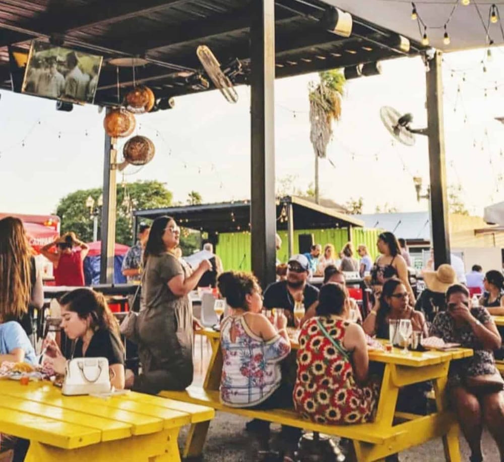 Outdoors social dining at QuestForDirections, vibrant setting with yellow tables, string lights, and diverse group enjoying food and drinks.