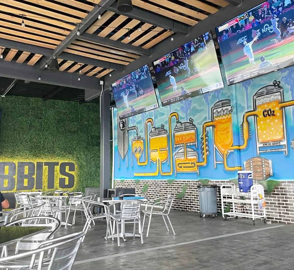 Outdoor bar with multiple TV screens showing a baseball game, modern decor, and beer brewing mural, ideal for sports fans.