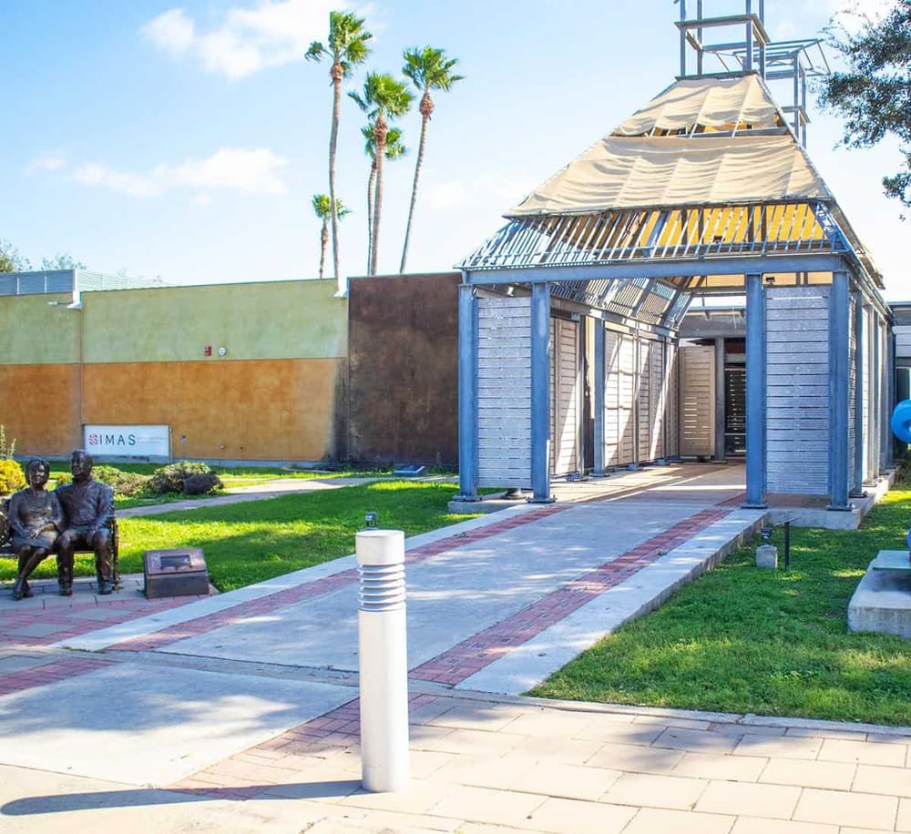 Outdoor event pavilion with blue wooden structure and modern statues in the park | Beautiful outdoor pavilion in a park setting, ideal for events and community gatherings | This image features a contemporary outdoor pavilion built with blue wood, situated in a lush green park with statues, palm trees, and a sunny sky, showcasing a perfect space for outdoor events and community activities.
